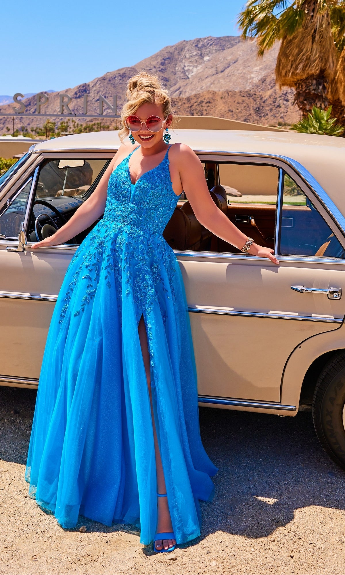 A woman in a Glitter-Tulle Long Ellie Wilde Prom Dress EW34089 stands before a beige vintage car in a sunlit desert landscape.